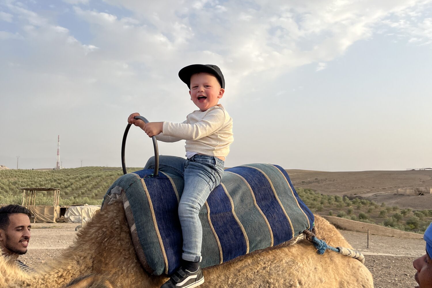 A child wearing a black cap and jeans, joyfully riding a camel with a blue striped saddle at a camp in Agafay.